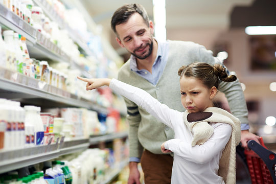 Family grocery shopping in supermarket: cute little girl pouting and pointing at milk bottle at dairy aisle with dad