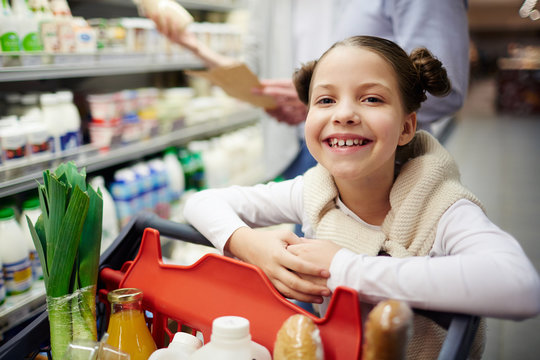 Portrait of cute little girl smiling happily leaning on shopping cart while buying groceries in supermarket with family