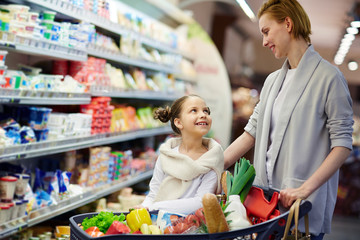 Portrait of happy woman shopping in grocery store with little girl, smiling and looking at each other caringly while pushing cart full of food