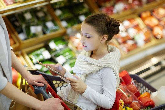 Portrait of little girl sitting in shopping cart and entertaining herself with digital tablet while her parents grocery shopping in supermarket