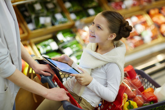 Portrait of smiling little girl sitting in shopping cart and entertaining herself with digital tablet while her parents grocery shopping in supermarket