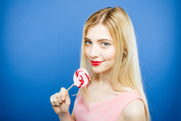 Pretty Blonde with Red Sensual Lips and Wonderful Smile is Holding Sweet Lollipop near Her Face. Portrait of Amazing Girl in Pink Dress on Blue Background in Studio.
