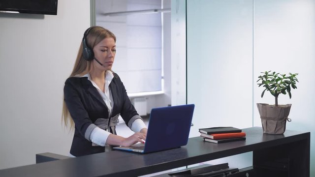 Sale manager talking with customer by phone using headset typing on computer. Happy woman with blond hair check information or place an order and delivery for client.