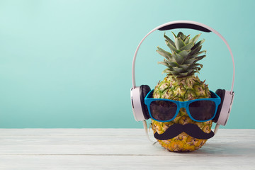 Pineapple with sunglasses and headphones on wooden table over mint background. Tropical summer...