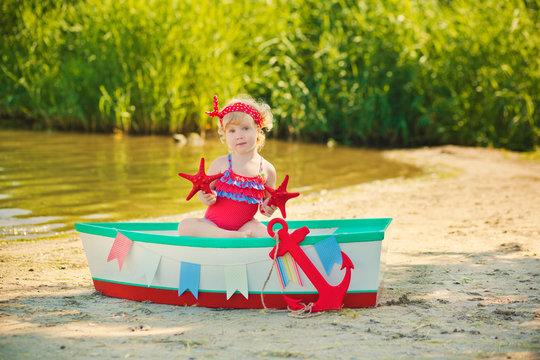 A cute little girl in a red bathing suit standing on the beach against the backdrop of boats