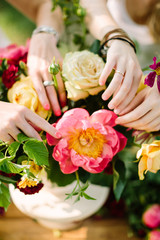 bouquet, people and floral arrangement concept - close-up on gentle hands of two young women decorating elegant bouquet of fresh summer yellow and white roses, pink peonies, red carnations and daisies
