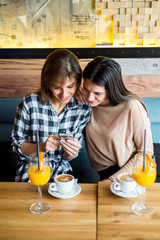 Two young female friends sitting in cafe, drinking coffee and juice, watch something on a mobile phone