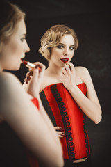 Beautiful young woman stands in front of a mirror and paints her lips. Retro (vintage) style