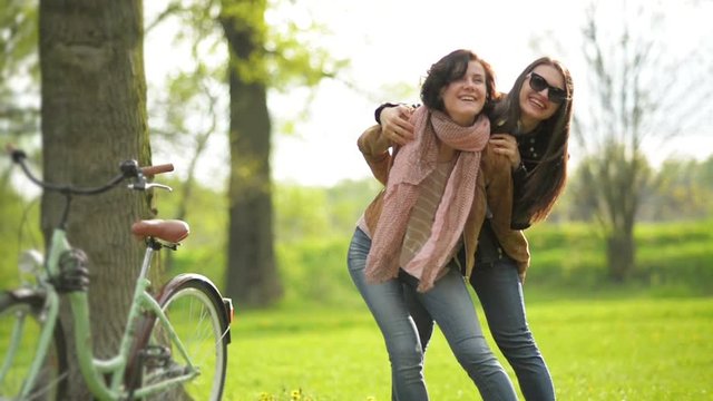 Pretty Woman with Short Curly Hair Wearing Jacker and Jeans is Carrying Her Female Friend in Sunglasses on the Shoulders. Smiling Happy Girls Spending Time Together Outdoors in the Park.