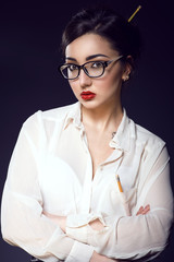 Young beautiful sexy business lady with updo hair wearing white silk blouse and trendy glasses in modern frame. There is a pencil in her breast pocket and another one in her hair. Her arms crossed