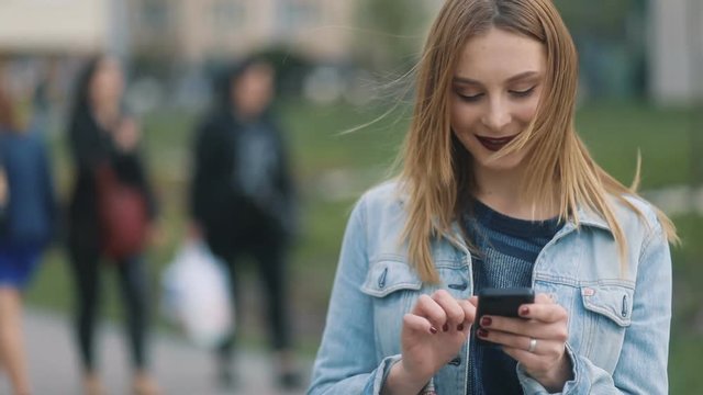 Amazing happy girl with dark-red lips girl in bluedenim court and strict bluse and blonde hair using her mobile smartphone and smiling in the urban city background Steadicam slow motion shot