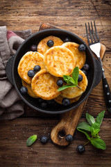 Delicious cottage cheese pancakes or syrniki with fresh blueberry in cast-iron pan on dark wooden rustic background, above view. Tasty breakfast.