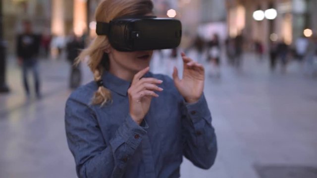 Handheld shot of surprised and excited young blonde woman with wireless portable VR headset goes through her first experience with virtual reality on urban street in city center at twilight time.