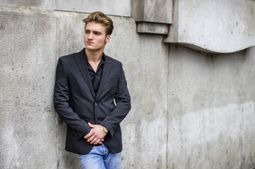Obraz premium Handsome blond young man, blue eyes, leaning against white, rough stone wall, looking up to a side