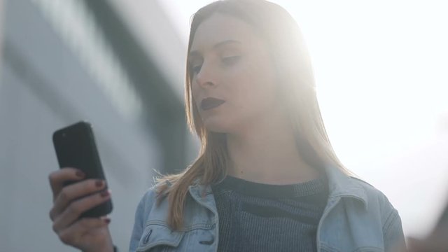 Sumptous european girl similar to famous actress with dark-red lips in blue denim court and strict bluse lordly communicating with someone, sun shining, urban street background