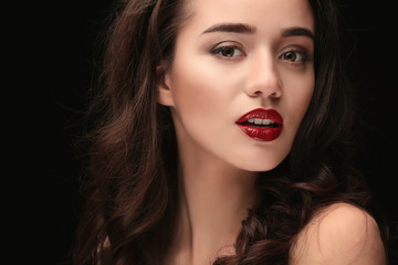 Fototapeta premium Young woman with glossy red lipstick and wavy brown hair on dark background, beauty and makeup