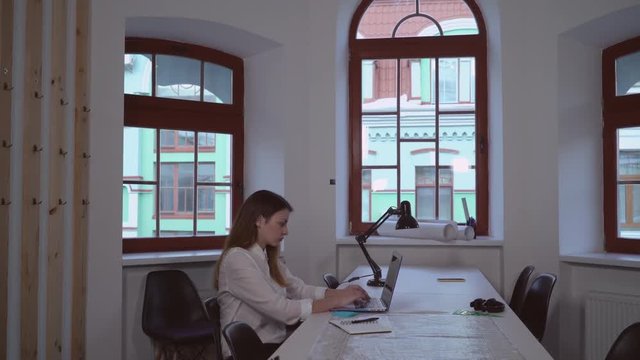 Young designer in architect agency typing on computer. Caucasian worker sitting at the working place on desk plan and blueprint.
