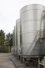Row of Large Stainless Steel Commercial Wine Holding Fermentation Tanks, Cement Ground, Outdoors, Daytime - Oregon