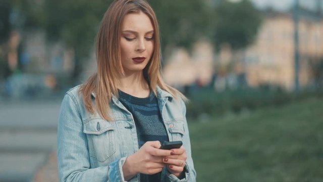 Glorious young woman similar with dark-red lips in blue denim court tapping on her smartphone with serious face, sunny weather, urban city background, chloe moetz, chloe, sun, shining, smile, smiling