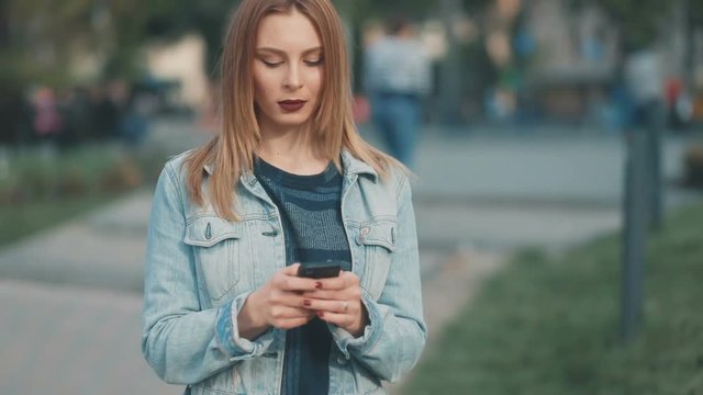 Young gentle girl, with european facial features with dark-red lips in blue denim court and strict bluse staying in the middle of the park lane and using her smartphone Steadicam slow motion shot