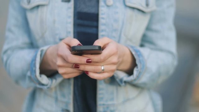 Extreme closeup of hands of young beatiful girl with dark-red manicure in blue denim court and strict bluse staying on the urban street and using her smartphone Steadicam slow motion shot