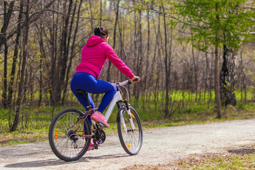 Fototapeta na wymiar Sporty young woman in a bright pink jacket and jeans rides a bike through the forest on a summer day. Back view