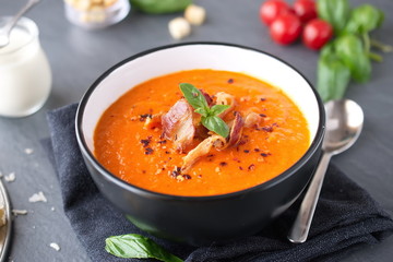 Thick tomato soup with basil and fried bacon in a black ceramic bowl on a grey abstract background....