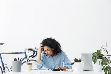 Serious dark-skinned woman accountant wearing formal shirt and eyeglasses calculating bills,...