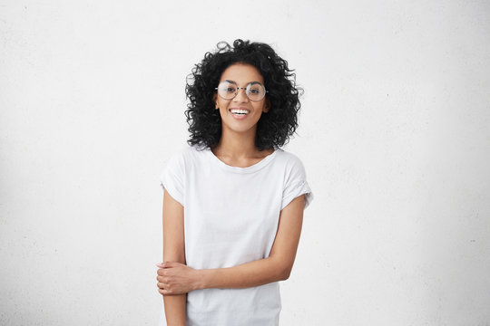 Indoor portrait of charming young mixed race female model dressed casually feeling shy and awkward during her first photo shoot, standing at white blank studio wall with copy space for content