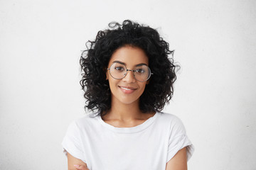 Fototapeta Close up studio shot of beautiful young mixed race woman model with curly dark hair looking at camera with charming cute smile while posing against white blank copy space wall for your content obraz