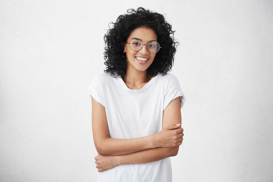 Human emotions and feelings. People, youth and happiness. Indoor shot of charming young mixed race woman model smiling broadly with her white straight teeth, holding arms crossed posing in studio
