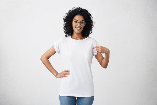 Clothing, design and advertising concept. Indoor shot of positive friendly young mixed race female in glasses pointing at copy space on her blank white t-shirt for your text or promotional content