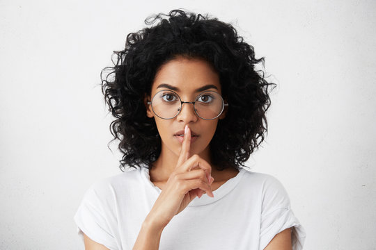 Worried serious dark-skinned girl wearing casual t-shirt and stylish round spectacles holding index finger at her lips, asking to keep silent and not to make noize. Human facial expressions