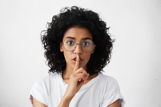 Don't tell anyone. Indoor shot of attractive serious dark-skinned girl pressing finger against her lips, saying Shh, Hush, demanding keeping secret and holding tongue, looking worried . Body language