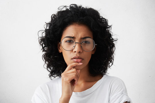 Let me see. Beautiful young woman in white t-shirt and big round spectacles squeezing her eyes in pensive expression, touching her chin while making choice or taking serious important decision