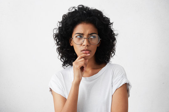 Hmm. Suspicious thoughtful young mixed race female with black curly hair looking up, keeping hand on her face as if trying hard to remember something important. Human face expressions and emotions