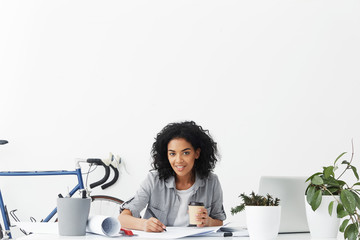 Job and career. Profession and occupation. Cheerful young female trainee holding paper cup of takeaway coffee while working on her first engineering project, looking ah camera with happy smile