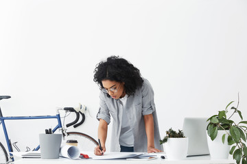 Attractive stylish mixed race female chief engineer in round glasses standing over her white desk and making drawings while finishing work on new engineering project, looking focused and serious