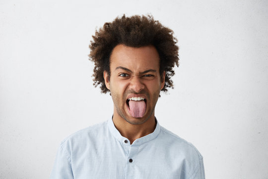 Emotional and goofy young African American man employee wearing blue shirt grimacing, making mouths, sticking out his tongue at camera trying to tease someone, acting like a little naughty child