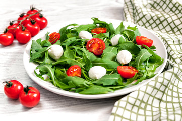 Vegetarian food and healthy lifestyle concept - spring salad with cherry tomato, mozzarella cheese and arugula on a white plate laying on wooden table. Branch of tomatoes and napkin with fork nearby.