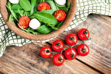 Branch of organic ripe fresh cherry tomatoes and wooden bowl with vegetarian healthy salad with tomato, mozzarella cheese and arugula laying on old vintage wooden texture background. Top view, flatlay