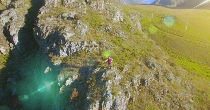Aerial UHD 4K view. Flight from (flight to) young hiking man standing on top of the cliff at mountain meadow and rural road at sunny summer day