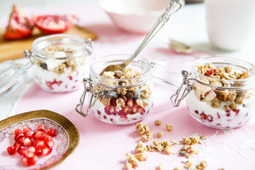 Obraz na płótnie Canvas Diet dessert with yogurt, granola and pomegranates in a glassware. Three jars. Horizontal. Rose tablecloth, silver spoon, wooden board. Closeup.