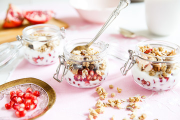 Diet dessert with yogurt, granola and pomegranates in a glassware. Three jars. Horizontal. Rose tablecloth, silver spoon, wooden board. Closeup.