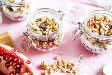 Diet dessert with yogurt, granola and pomegranates in a glassware. Three jars. Top view. Rose tablecloth, silver spoon, wooden board. Closeup.