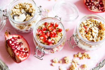 Diet dessert with yogurt, granola and pomegranates in a glassware. Three jars. Top view. Rose tablecloth, silver spoon, wooden board. Closeup.