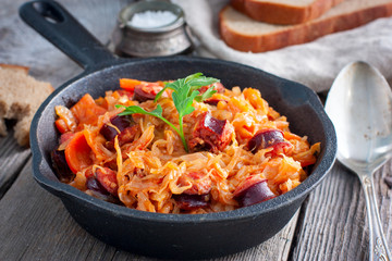 Stewed cabbage with sausages in a portioned cast-iron frying pan on a wooden table, horizontal