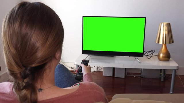 Girl Uses Remote Control of Television Green Screen. Young Woman watching television with green screen, shot behind models shoulders