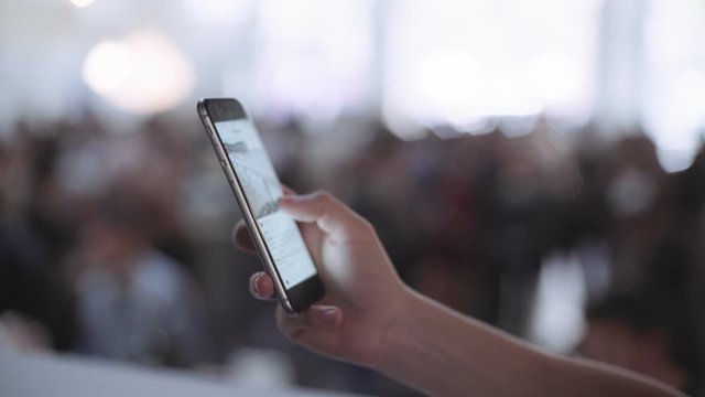 Close up of a female hand scrolling through applications on her smartphone, with unfocused and blurry party with lot of people in background