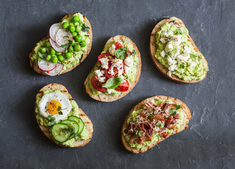 Variation avocado sandwich - with crispy bacon, quail egg, tomatoes, goat cheese, green peas, radish, cucumber. Healthy snack. On a dark background, top view